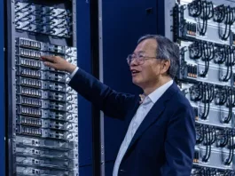 A man in a suit stands in a server room, reaching towards a rack of Supermicro computer servers with a smile on his face. Computer equipment and cables are visible in the background, echoing recent news about tech industry developments.